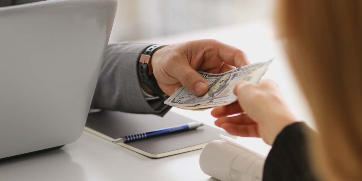 A hand behind a computer is handing money to a consultant they are working with.
